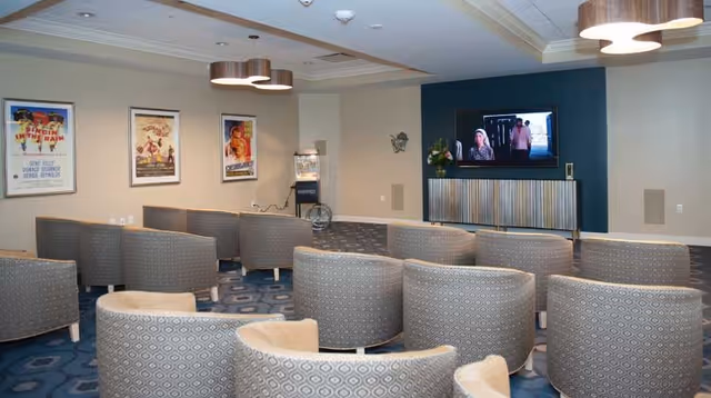 A cozy media room with multiple rows of cushioned chairs facing a wall-mounted television. The room has a patterned blue carpet, framed movie posters on one wall, and modern ceiling lights. There is also a popcorn machine in the corner.