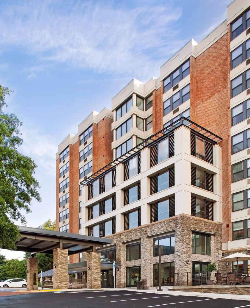 Multi-story brick and stone senior living building with a covered entrance and large windows under a blue sky.