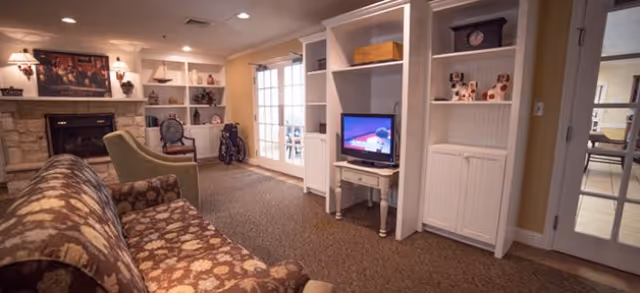 A cozy living room area with a floral patterned sofa, a green armchair, a white entertainment center with a TV, and a stone fireplace. The room has beige walls, carpeted floor, and French doors leading to another room. Various decorative items and a wheelchair are visible in the background.