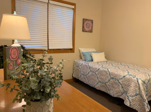 A cozy single bedroom with a twin bed covered by a blue-and-white quilt, a wooden table with a lamp and potted plant, and a window with blinds.