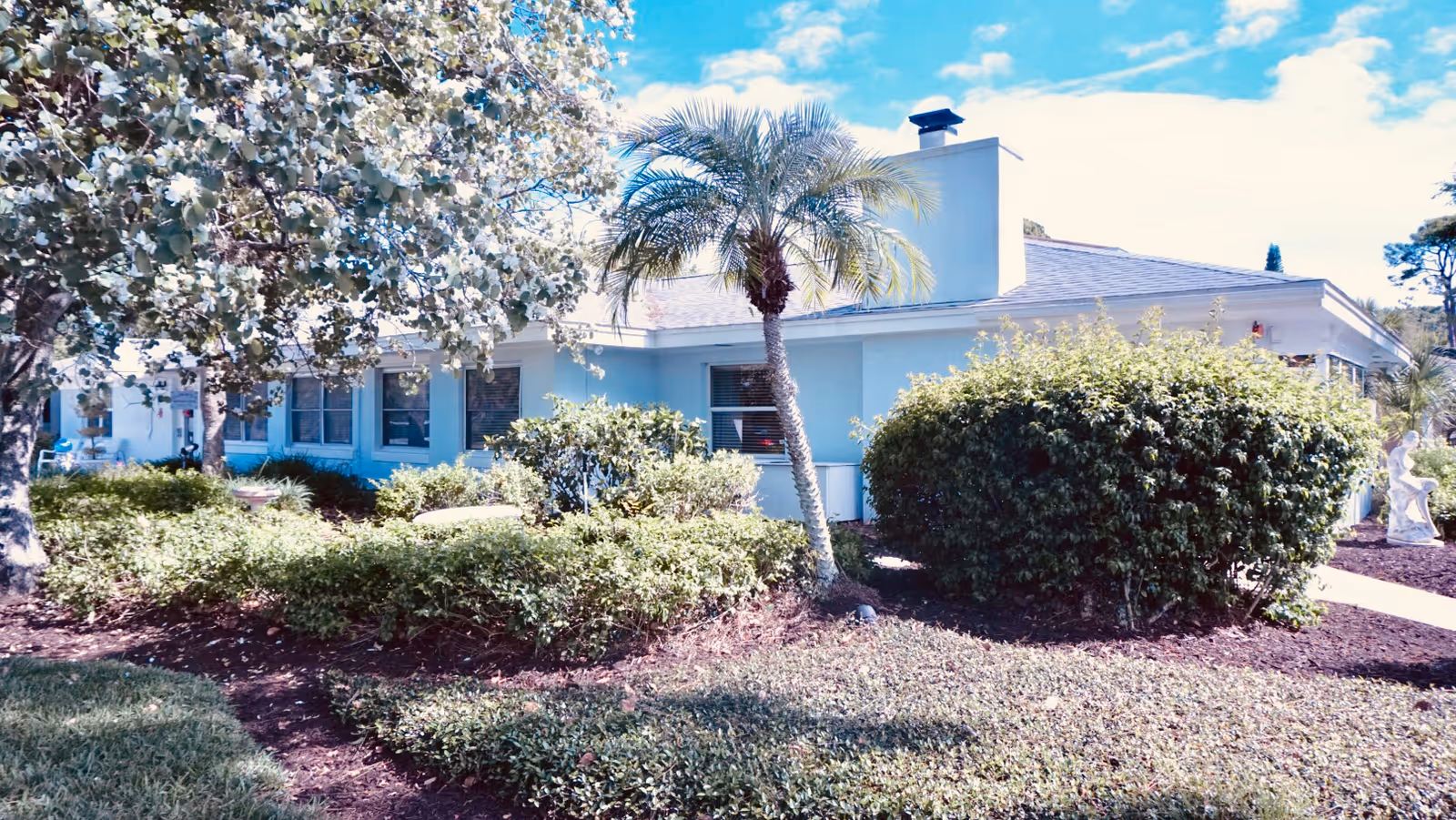 Exterior view of a single-story assisted living facility building surrounded by well-maintained landscaping including bushes, a palm tree, and other greenery under a partly cloudy sky.