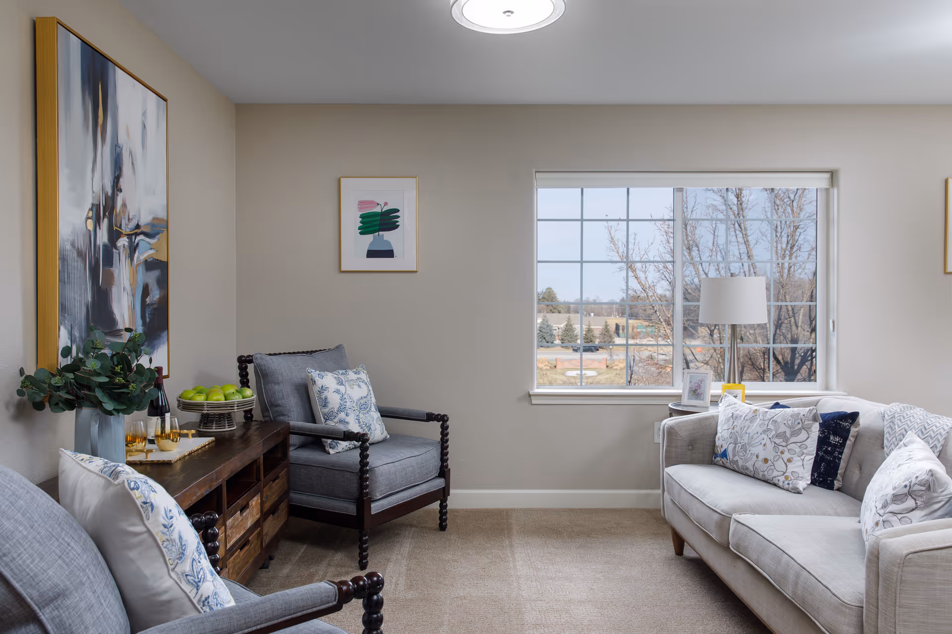A cozy living room with a large window showing an outdoor view. The room features a light gray sofa with patterned pillows, two gray armchairs with wooden frames and patterned cushions, a wooden sideboard with green apples and glasses on a tray, a floor lamp near the window, and two framed artworks on the walls.