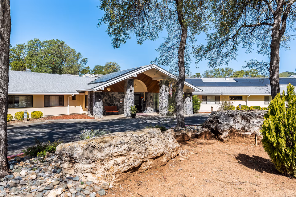 Exterior view of Meadowood Nursing Center building entrance with stone pillars, a covered porch, and surrounding trees and landscaping under a clear blue sky.