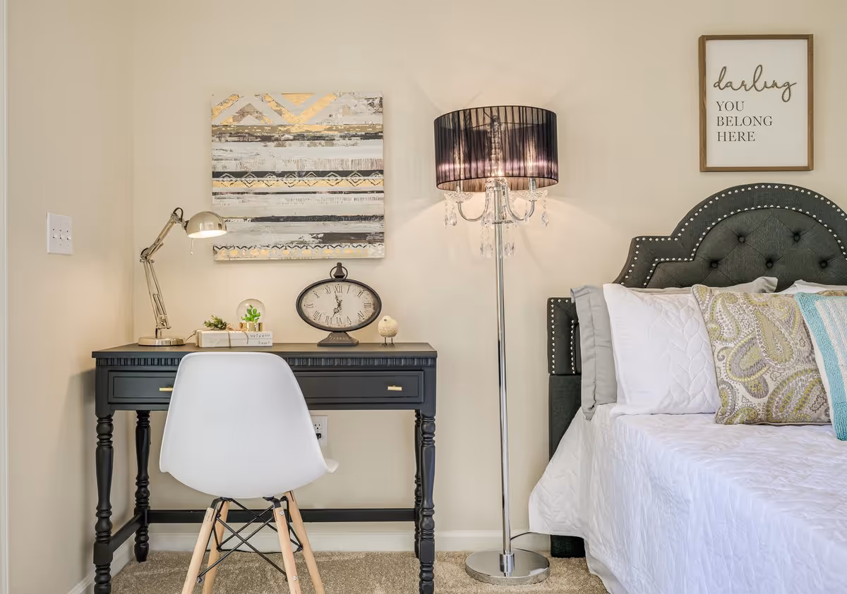 A cozy bedroom corner with a black writing desk and chair next to a tufted bed, decorative lamps, wall art, and pillows.