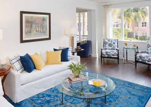 Bright and airy living room with a white sofa adorned with blue and yellow cushions, a glass coffee table with books and a decorative bowl, two patterned armchairs near a large window with a view of palm trees and a brick building, a blue patterned rug, and a framed painting on the wall.