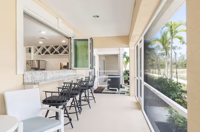 A bright and airy screened-in patio area with a row of black bar stools along a granite countertop serving window that opens to a kitchen. There is also a white chair with a cushion and a small round table. Outside the screened area, palm trees and greenery are visible.