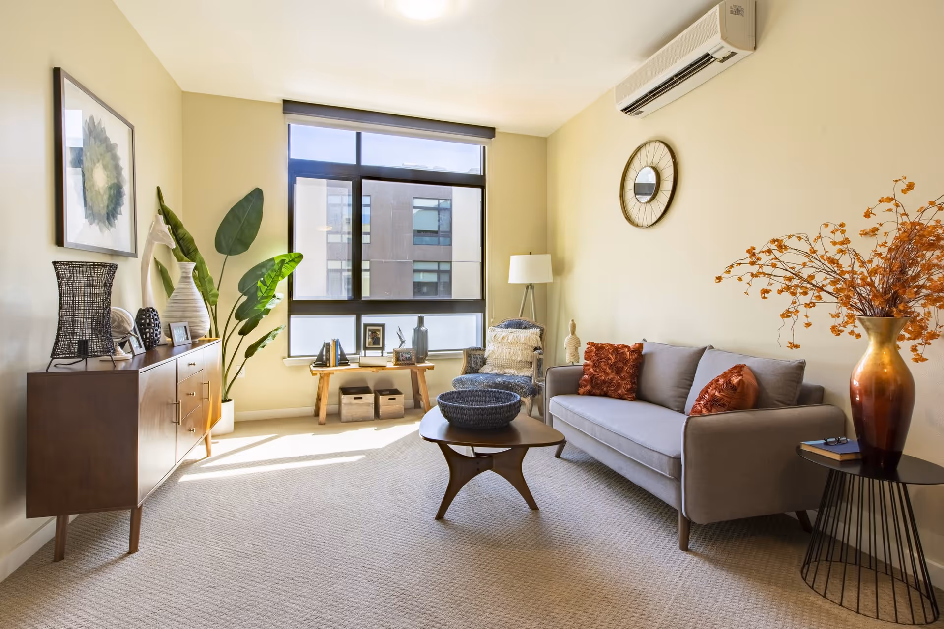 A bright and cozy living room with a large window letting in natural light. The room features a gray sofa with two orange decorative pillows, a wooden coffee table with a woven basket, a side table with a large vase of orange flowers, a floor lamp, a blue patterned armchair with a white throw, and a wooden sideboard with decorative items and framed pictures. A round wall clock hangs above the sofa, and a large green plant is placed near the window.