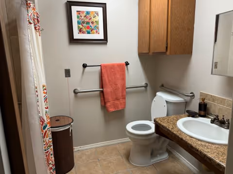 A bathroom with a white toilet, a sink with a granite countertop, a wooden cabinet above the toilet, a towel rack with an orange towel, a framed colorful geometric artwork on the wall, a shower curtain with a colorful pattern, and a brown laundry hamper.