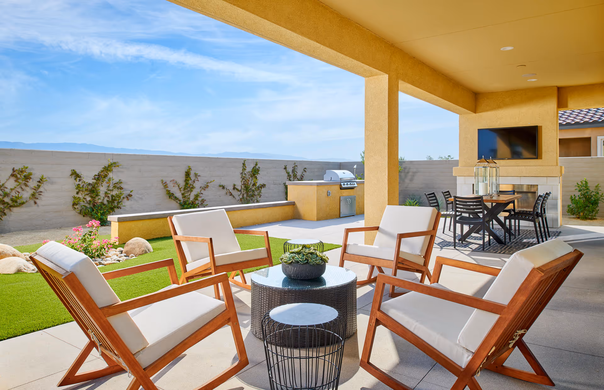 Covered outdoor patio area with four wooden chairs featuring white cushions arranged around a round glass-top table with a small plant centerpiece. In the background, there is a dining table with six black chairs, a built-in barbecue grill, and a mounted flat-screen TV above a fireplace. The patio overlooks a small garden with green grass, rocks, and plants, enclosed by a beige wall under a clear blue sky.