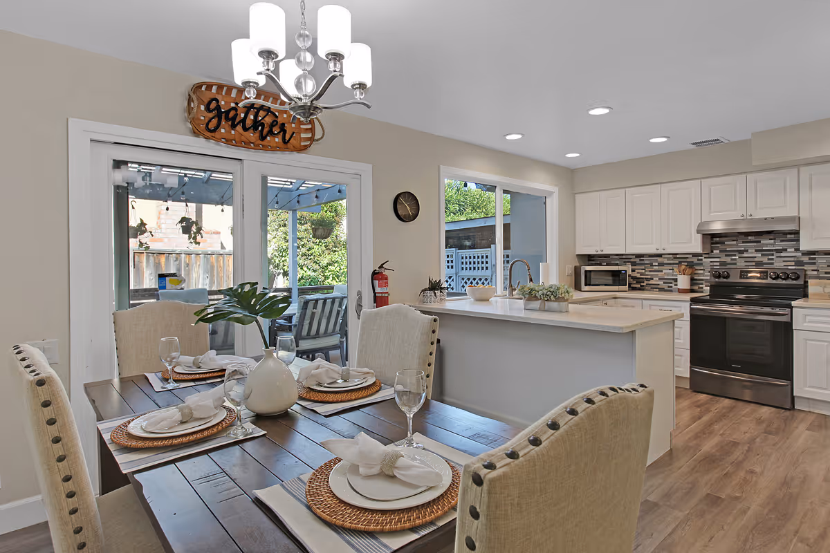 A bright and modern dining area connected to a kitchen. The dining table is set with four place settings, each with a white plate, napkin, and glass. A white vase with green leaves is centered on the table. The kitchen features white cabinets, a tiled backsplash, a black stove, microwave, and a kitchen island with a sink. Large windows and glass doors provide natural light and a view of an outdoor patio with hanging plants.