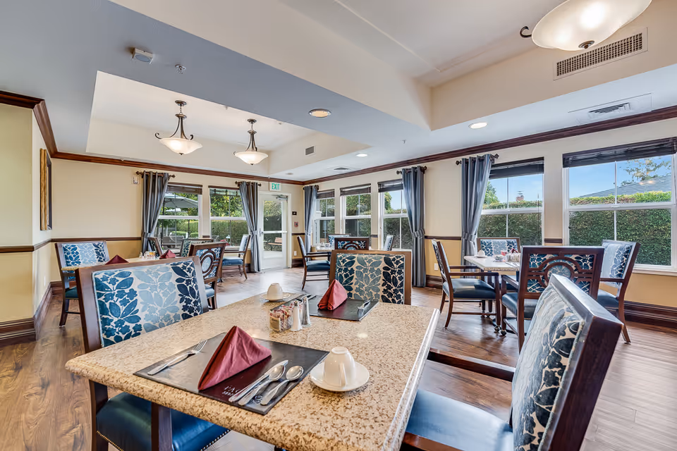 A bright dining room with several tables and chairs arranged neatly. Each table is set with utensils, napkins, and cups. Large windows with curtains allow natural light to fill the room, and there are two ceiling light fixtures. The room has wooden flooring and a door leading outside.