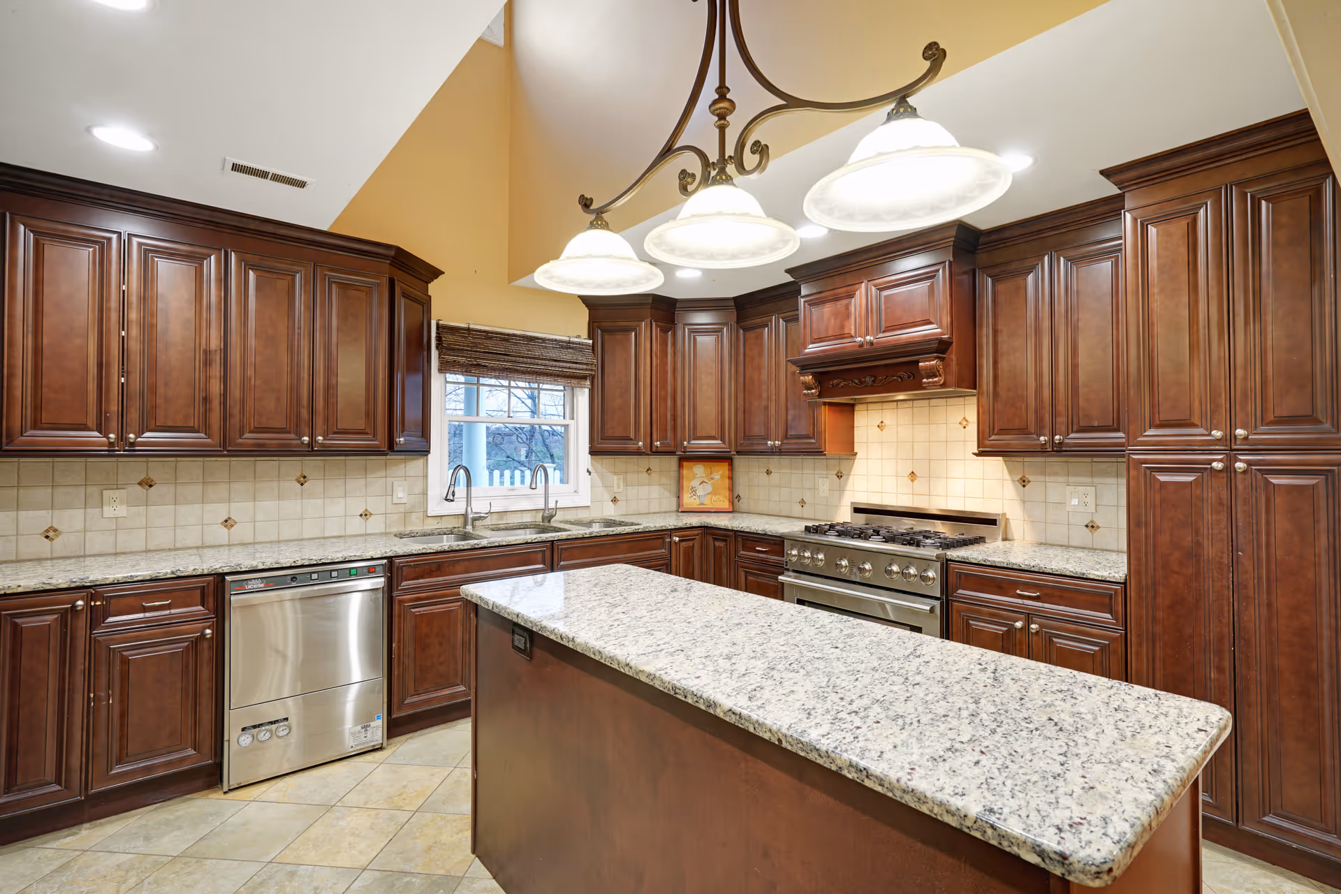 A spacious kitchen with dark wooden cabinets, granite countertops, a central island, a stainless steel dishwasher, and a large stove with an ornate wooden hood. The kitchen has tiled floors and a window above the sink letting in natural light. A chandelier with three lights hangs above the island.