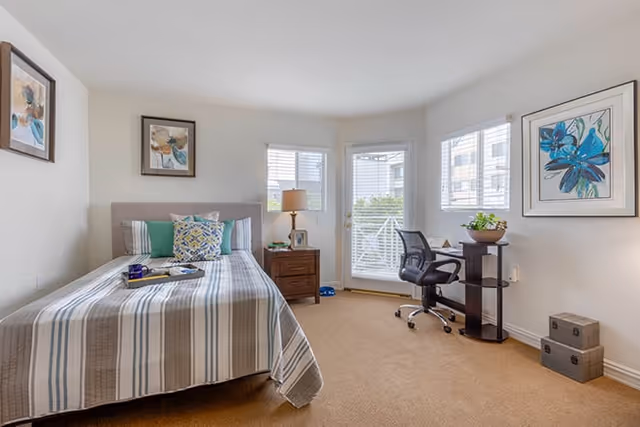 Bright bedroom with a striped bed, bedside table and lamp, a desk and office chair by windows and a glass door to a small balcony.