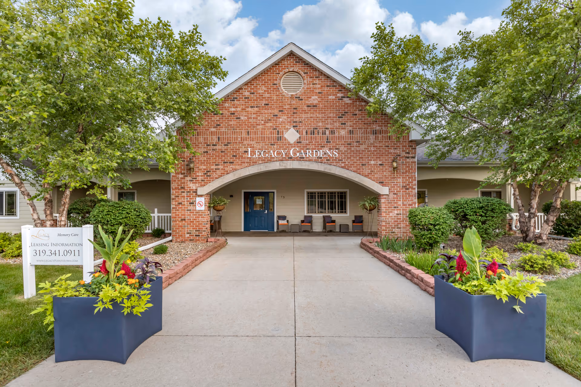 Front entrance of Legacy Gardens senior living facility with a brick archway, blue door, chairs on the porch, two large planters with green and red plants, and trees and shrubs on either side of the walkway.