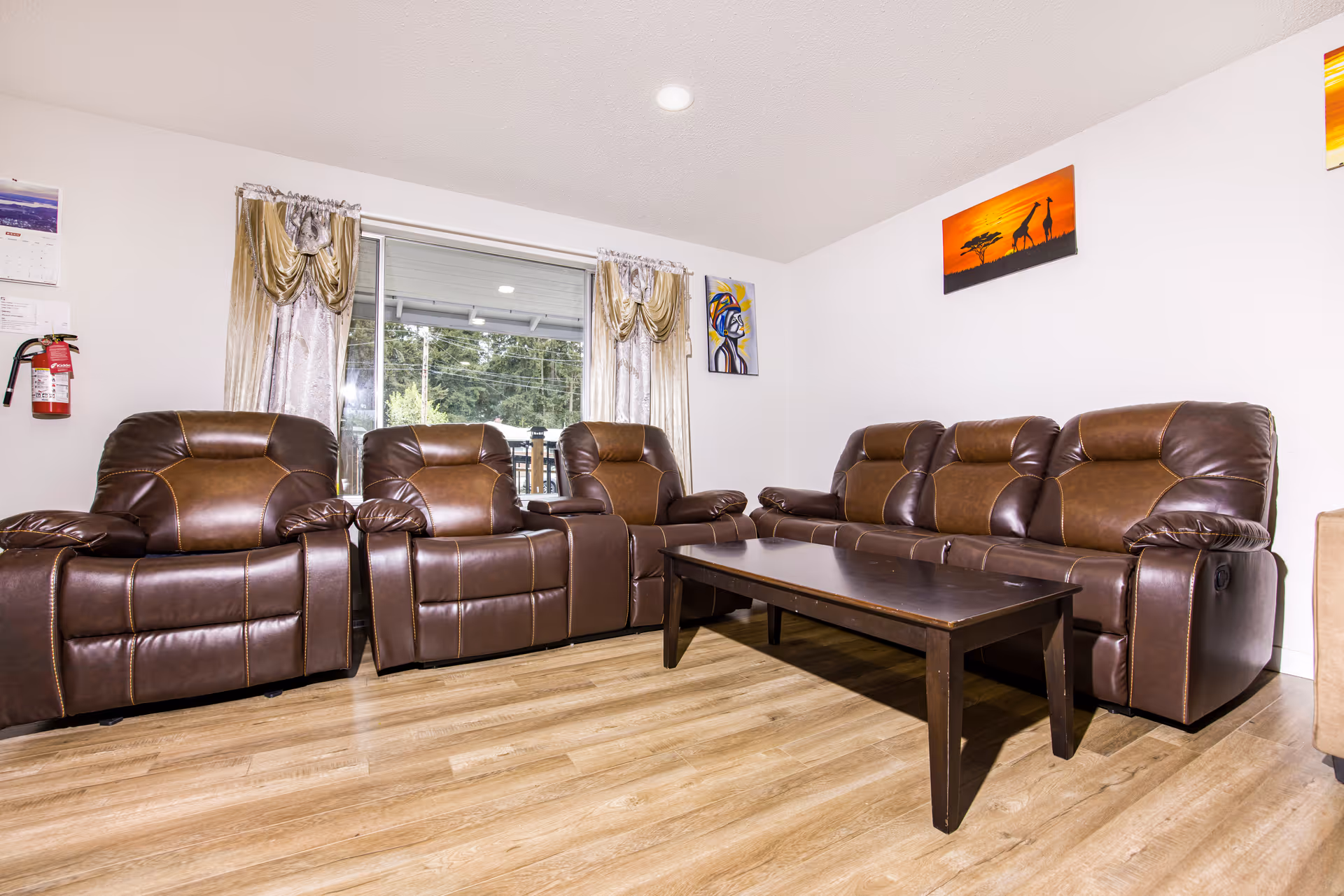 A living room with three brown leather recliners and a matching brown leather sofa arranged around a dark wooden coffee table. The room has light wood flooring, white walls, and a large window with gold curtains. There are colorful paintings on the walls and a fire extinguisher mounted near the window.