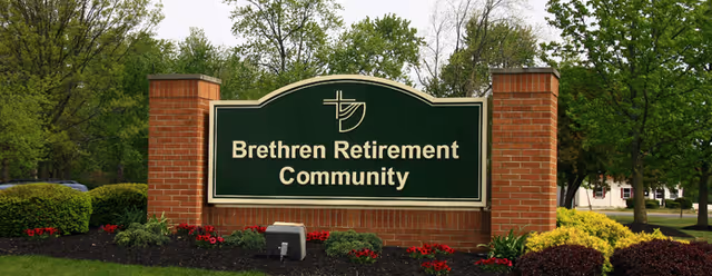 A large green and white sign reading 'Brethren Retirement Community' mounted on a brick base, surrounded by landscaped bushes, flowers, and trees in the background.