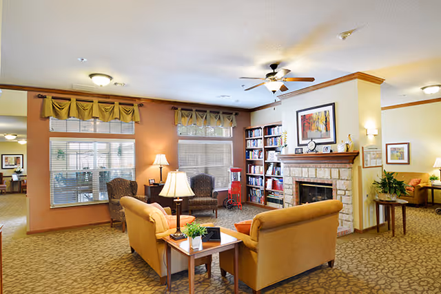 Cozy common room with sofas and armchairs arranged around a coffee table, a fireplace, bookshelves, and large windows.