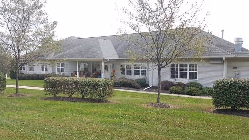 Single-story building with a gray roof and light-colored siding surrounded by green grass, small trees, and bushes. There is a sidewalk leading to the entrance with a covered porch area featuring outdoor seating.