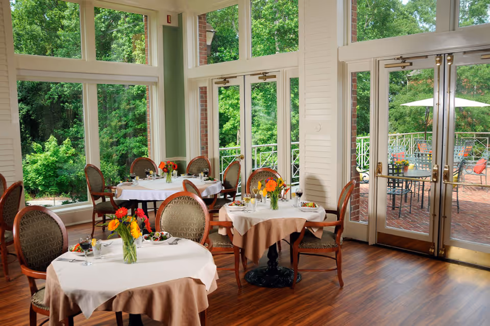 A bright dining room with round tables covered in white and beige tablecloths, each set with glasses, plates, and floral centerpieces. Large windows and glass doors provide a view of lush green trees and an outdoor patio with tables and umbrellas. The room has wooden flooring and elegant chairs with patterned upholstery.