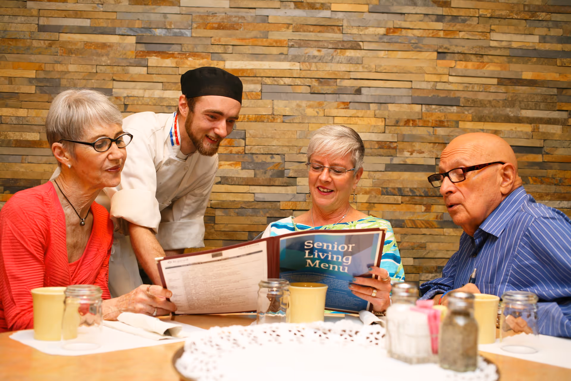 Three elderly people sitting at a dining table looking at a menu titled 'Senior Living Menu' with a chef standing beside them smiling and assisting. The background features a stone brick wall.