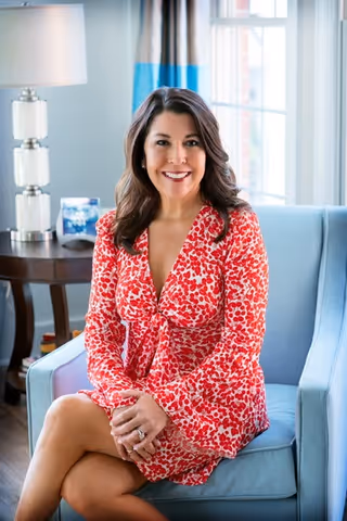 Smiling woman in a red patterned dress sitting cross-legged on a light blue armchair in a bright, well-lit living room.