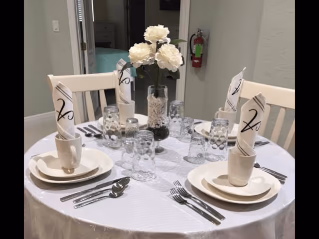 A round dining table set for four with white plates, cups, silverware, and napkins folded in an upright design. A glass vase with white flowers is placed in the center of the table. The background shows a hallway with a fire extinguisher mounted on the wall.