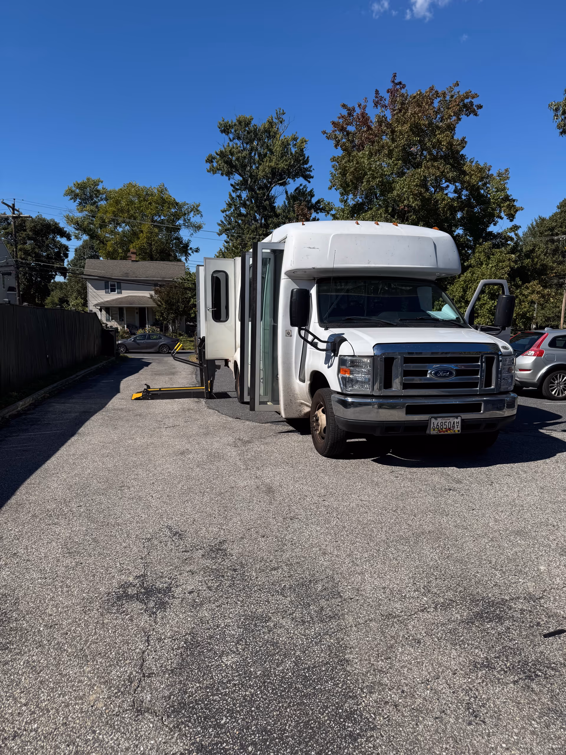 A white shuttle bus with its side door open and a wheelchair lift extended, parked on an asphalt driveway with trees and houses in the background under a clear blue sky.