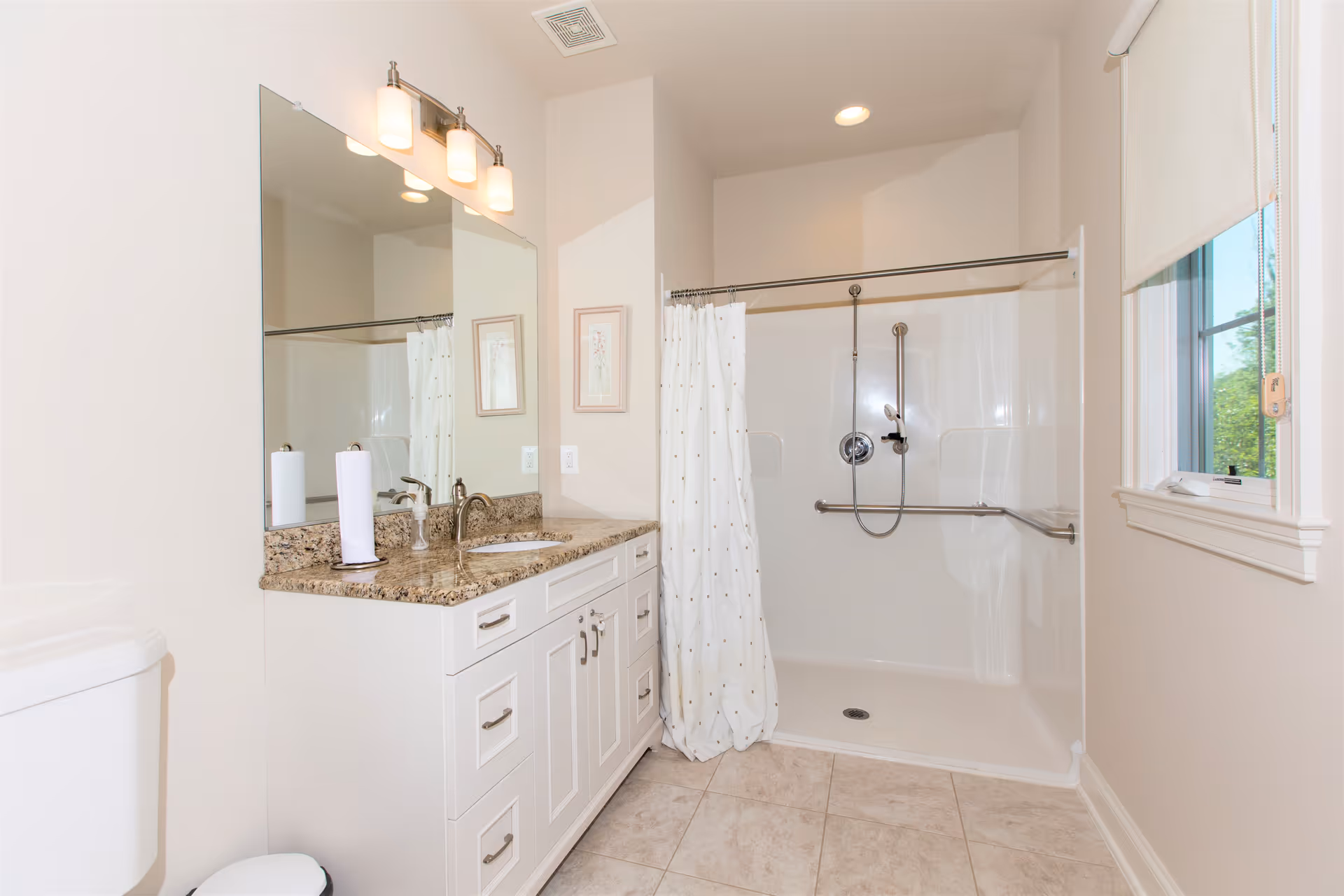 Bright bathroom with a large mirror above a granite countertop vanity with white cabinets, a toilet, and a walk-in shower with grab bars and a white shower curtain. There is a window with a white blind letting in natural light.