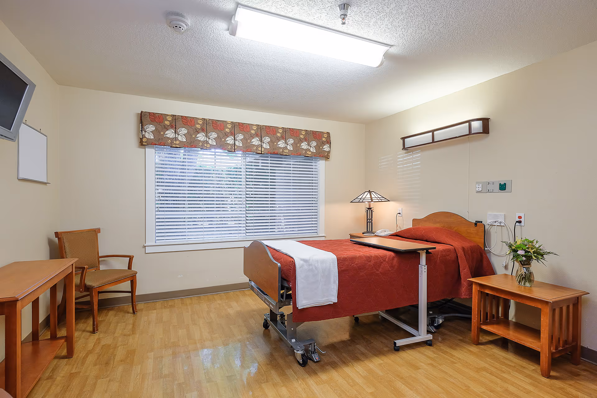 A clean and simple patient room with a hospital bed covered in a red blanket and a white towel, a wooden side table with a vase of flowers, a wooden chair, a small wooden table, a window with blinds and floral valance, and a ceiling light fixture.