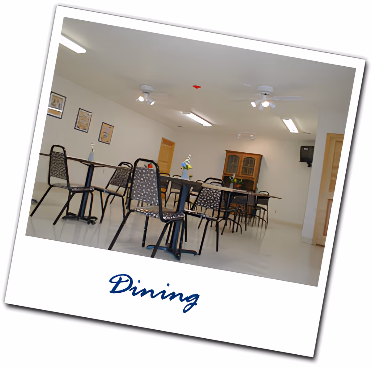 A dining room with several tables and chairs arranged neatly. The room has white walls with framed pictures, ceiling fans with lights, and a wooden cabinet against the far wall. There is a small TV mounted in the corner and a door on the right side.