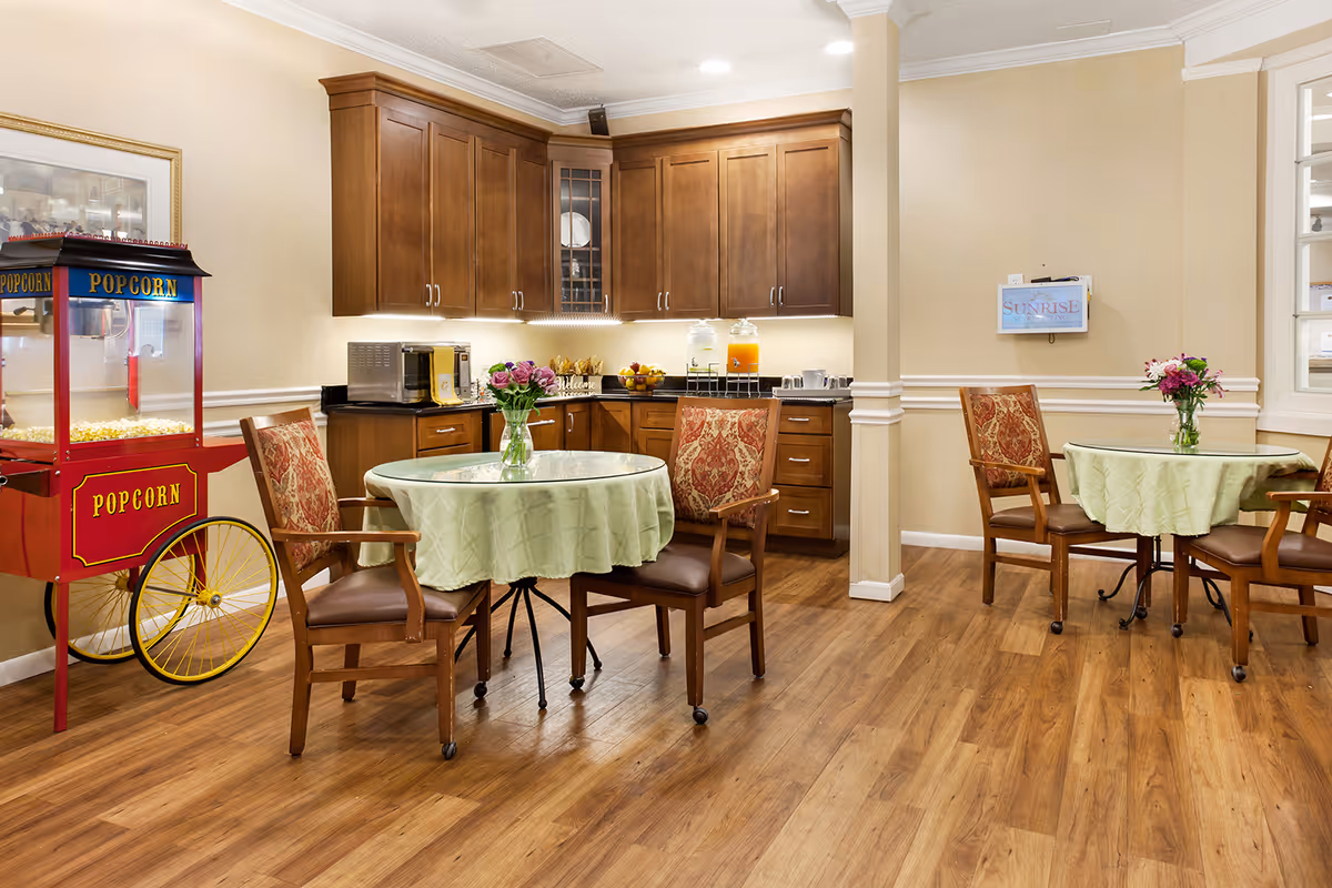 Communal dining area with round tables and chairs, a popcorn cart, and a kitchenette with wooden cabinets.