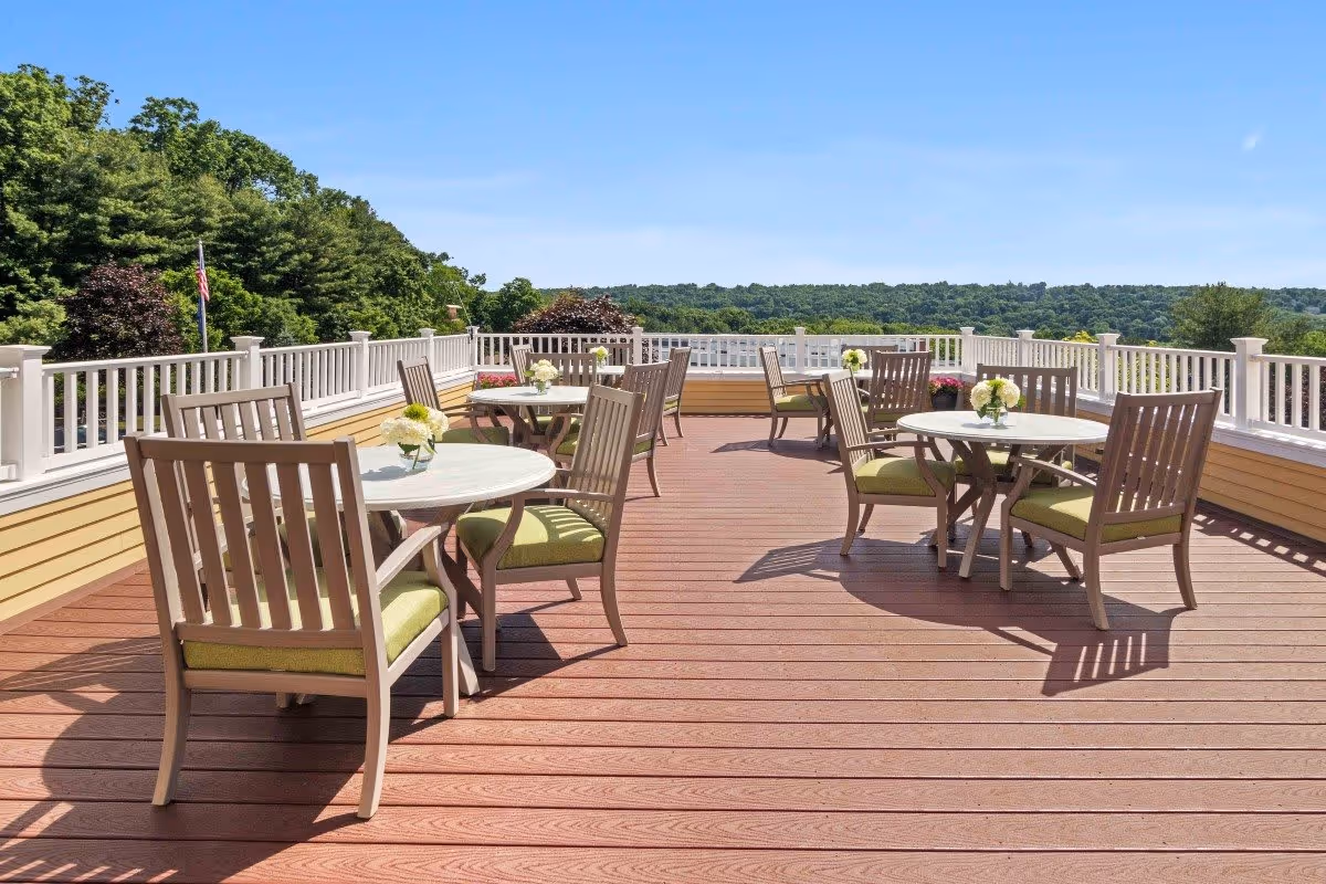 Outdoor patio area with several round tables and wooden chairs with green cushions, each table decorated with a small flower arrangement. The patio is surrounded by a white railing and overlooks a scenic view of trees and hills under a clear blue sky.