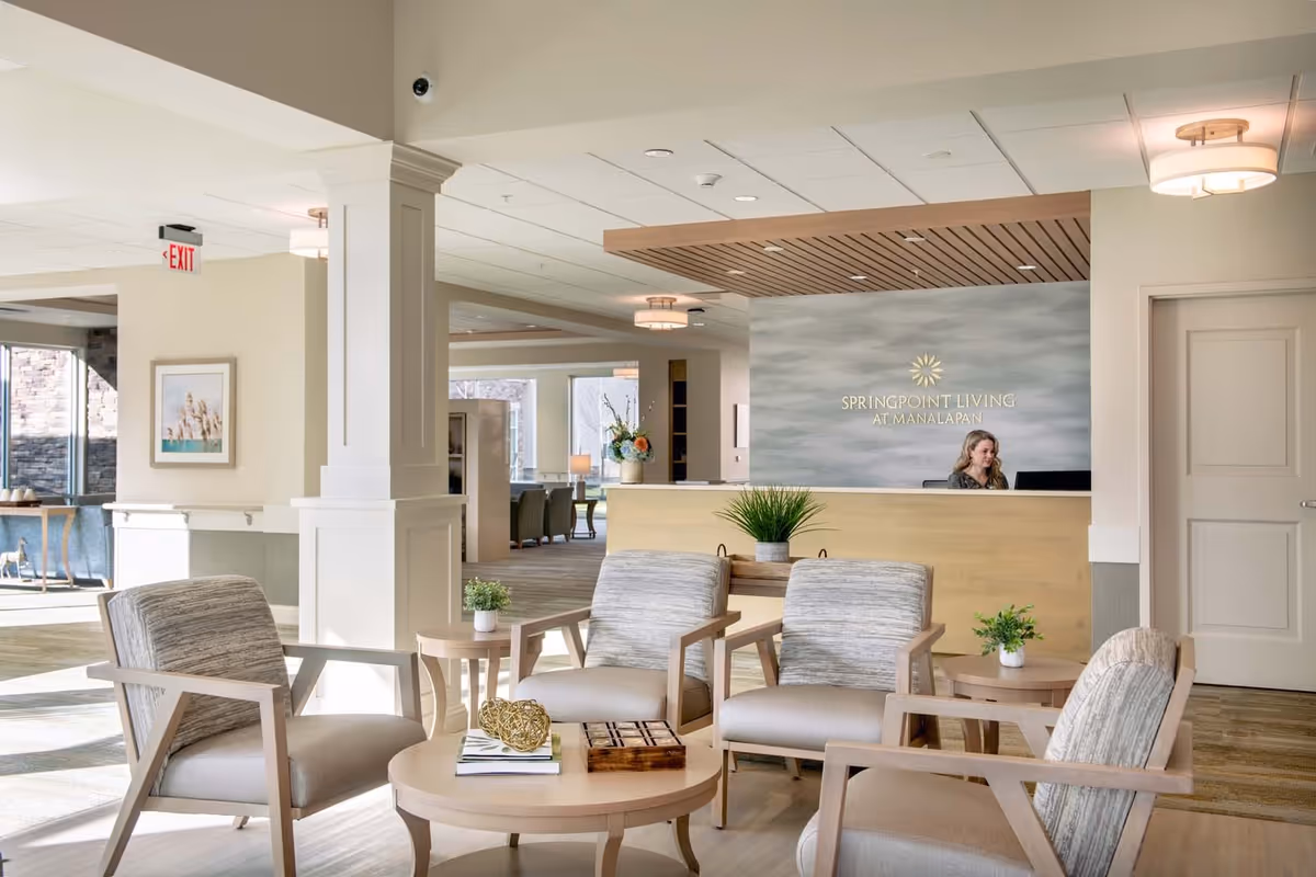 A bright and welcoming reception area of Springpoint Living at Manalapan featuring a receptionist behind a wooden desk with the facility's name on the wall. The seating area in the foreground has four cushioned armchairs arranged around a round wooden coffee table with decorative items and plants. The space is well-lit with natural light coming through large windows and modern ceiling lights.