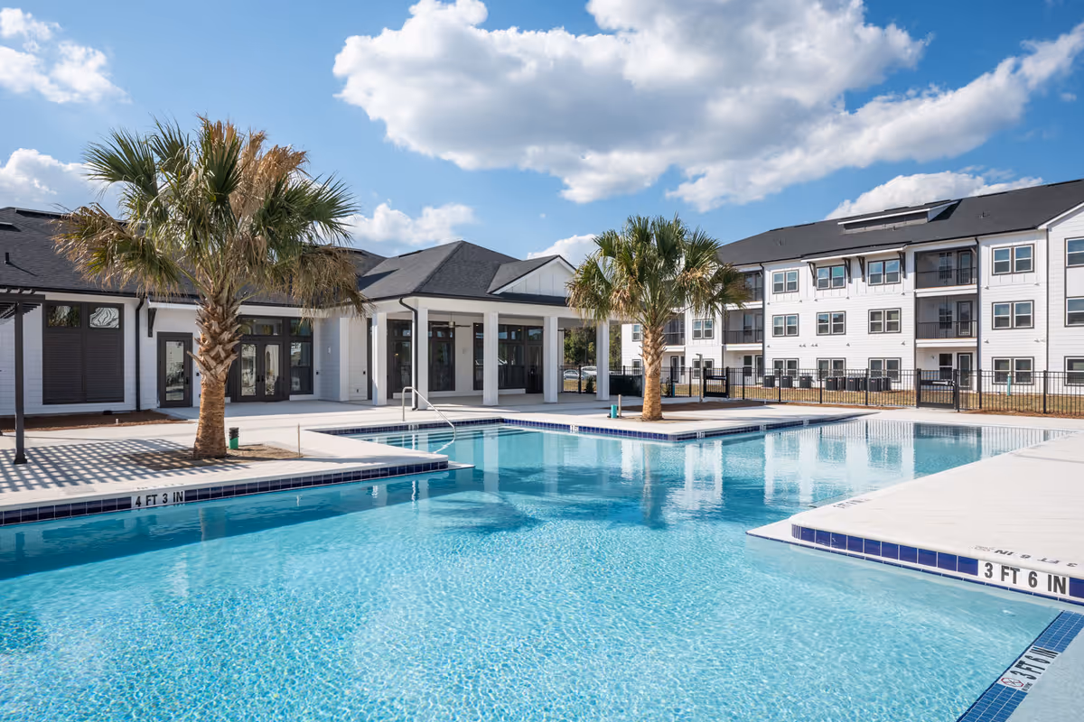 Outdoor swimming pool with clear blue water, surrounded by a concrete deck and two palm trees. In the background, there is a white multi-story building with many windows and a covered patio area under a partly cloudy blue sky.