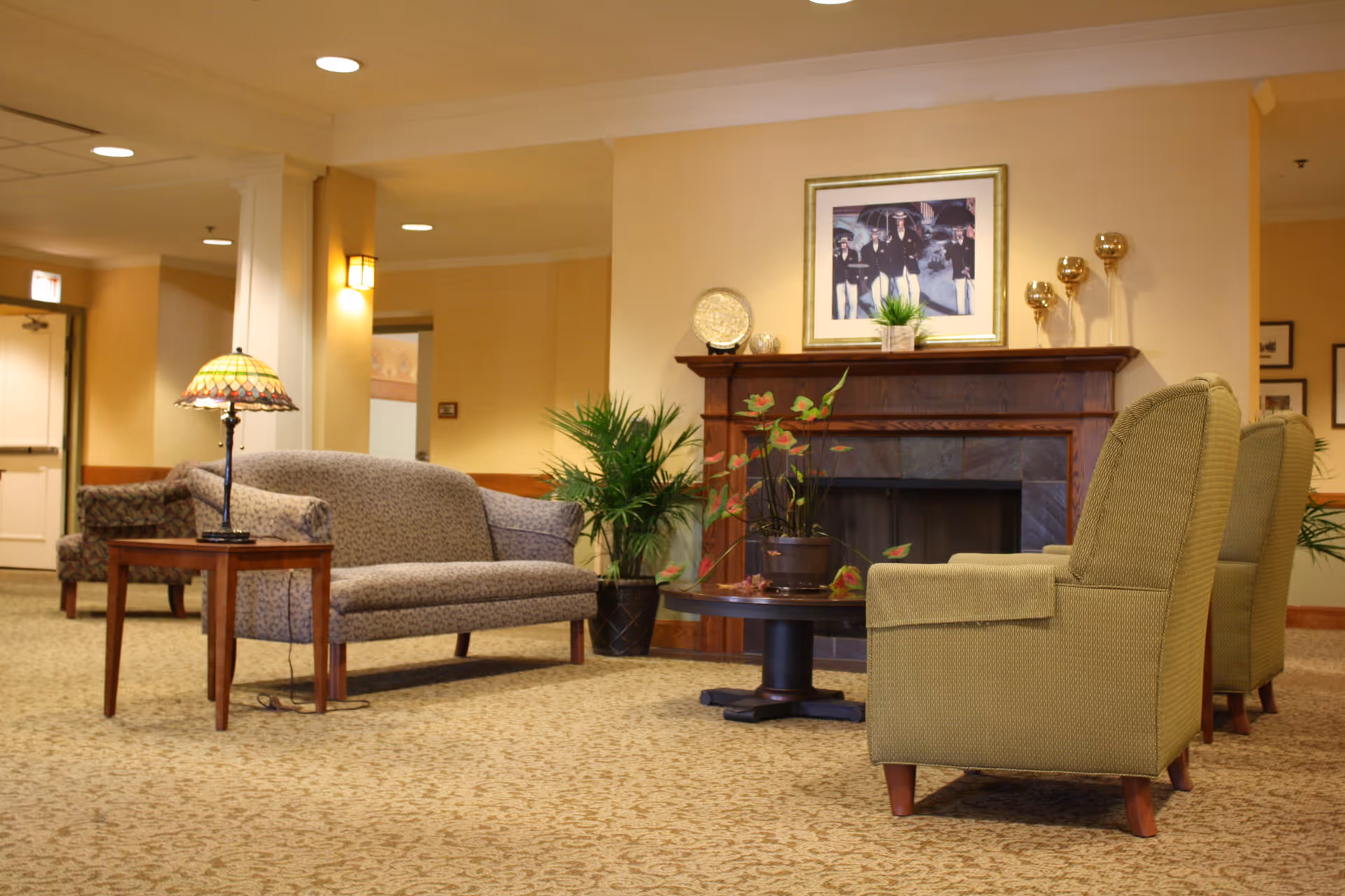 A cozy living room area in a senior living facility featuring a patterned loveseat, two green armchairs, a wooden coffee table with a potted plant, and a wooden side table with a Tiffany-style lamp. The room has a fireplace with decorative items and a framed picture above it. The walls are painted a warm beige color and the floor is carpeted with a patterned design.