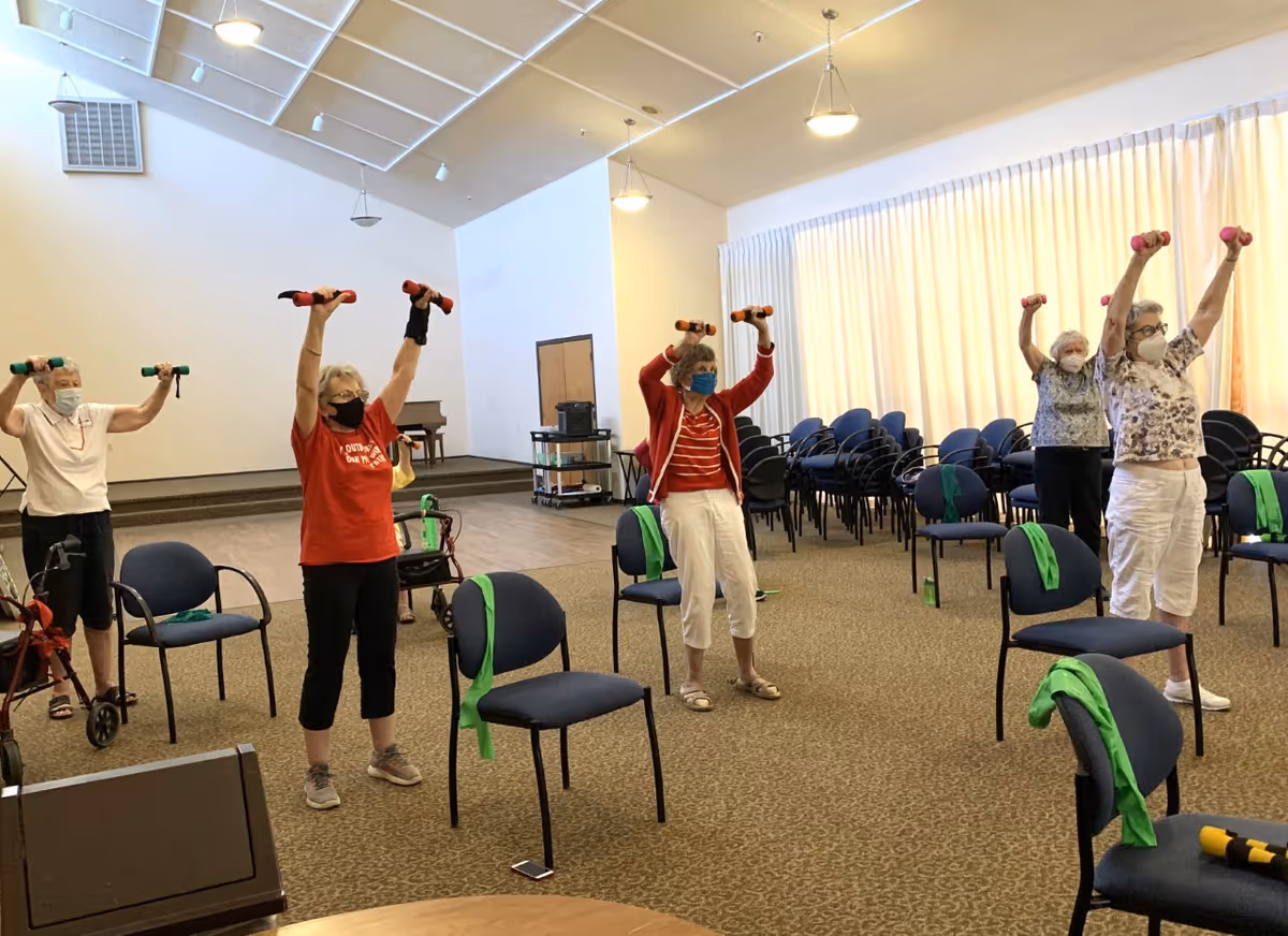 A group of elderly women wearing face masks participate in a seated exercise class in a spacious room. They are standing behind chairs, each holding small dumbbells raised above their heads. The room has carpeted floors, a stage area with a piano, and rows of stacked chairs along the walls.