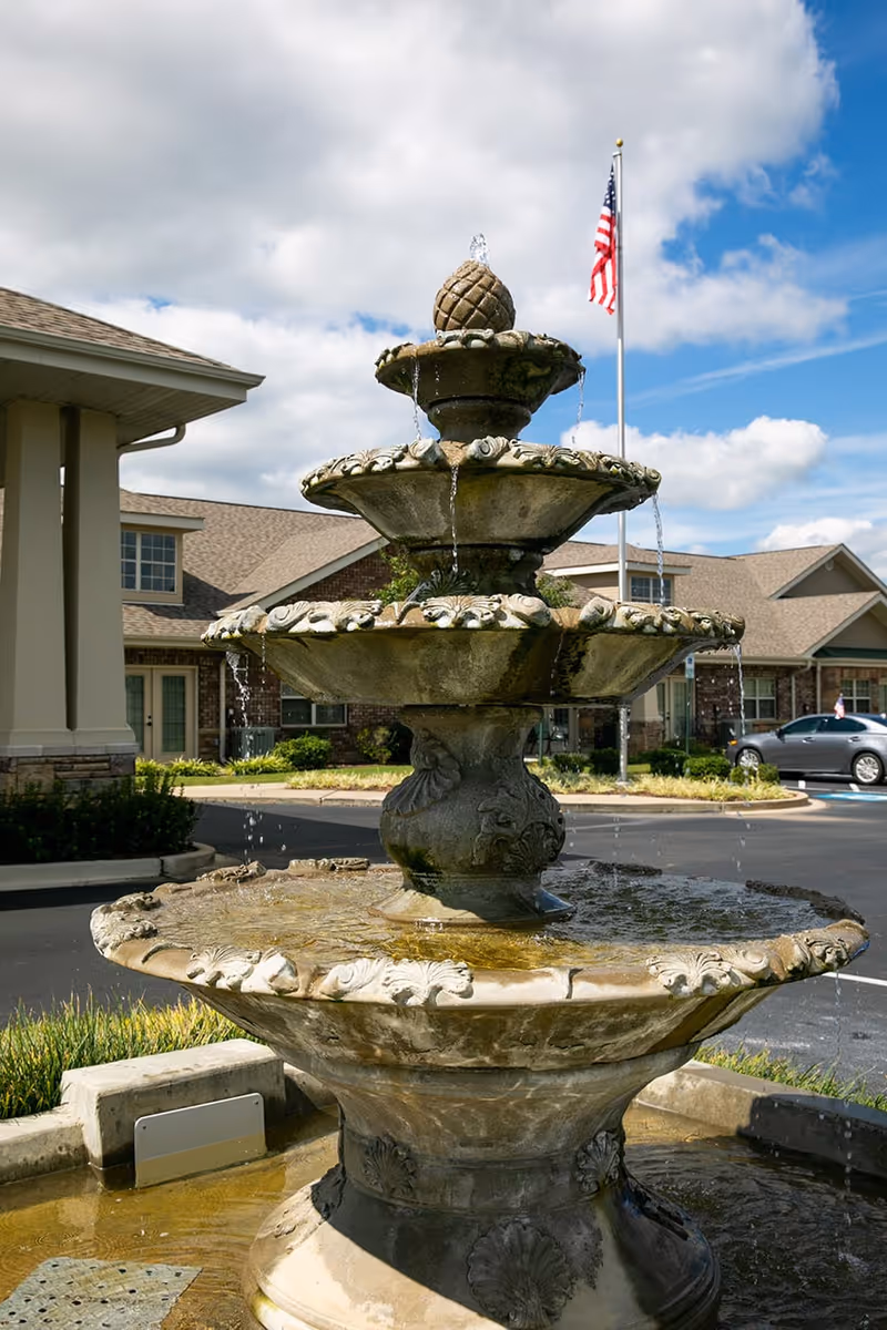 A multi-tiered stone water fountain with water flowing from the top tier to the lower basins, situated outdoors in front of a senior living facility building. An American flag on a flagpole is visible in the background under a partly cloudy sky.