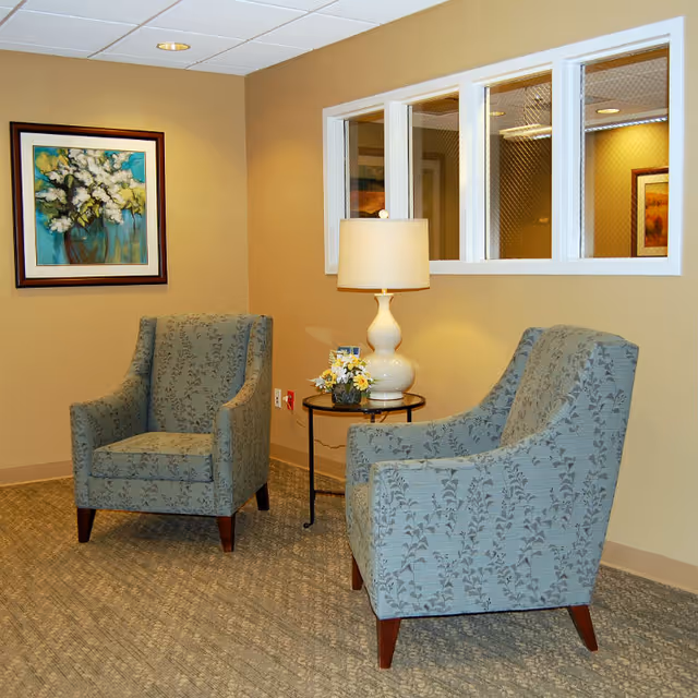 A cozy seating area with two patterned armchairs facing each other, a small round table between them holding a white table lamp and a small flower arrangement. The walls are painted beige, with a framed floral painting on one wall and three small windows with wire mesh on another.