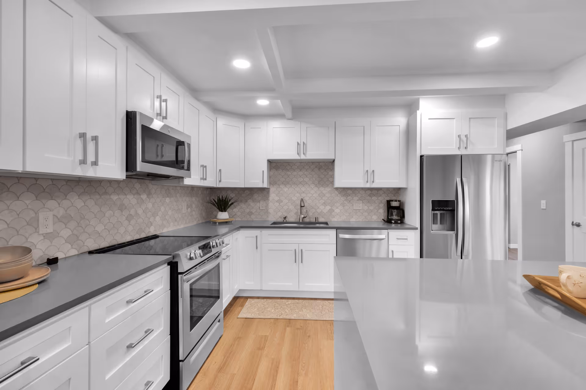Modern kitchen with white cabinetry, stainless steel appliances including a microwave, stove, dishwasher, and refrigerator. The kitchen features a light gray countertop, a decorative tile backsplash, recessed ceiling lights, and a wooden floor. A small plant and coffee maker are placed on the countertop.