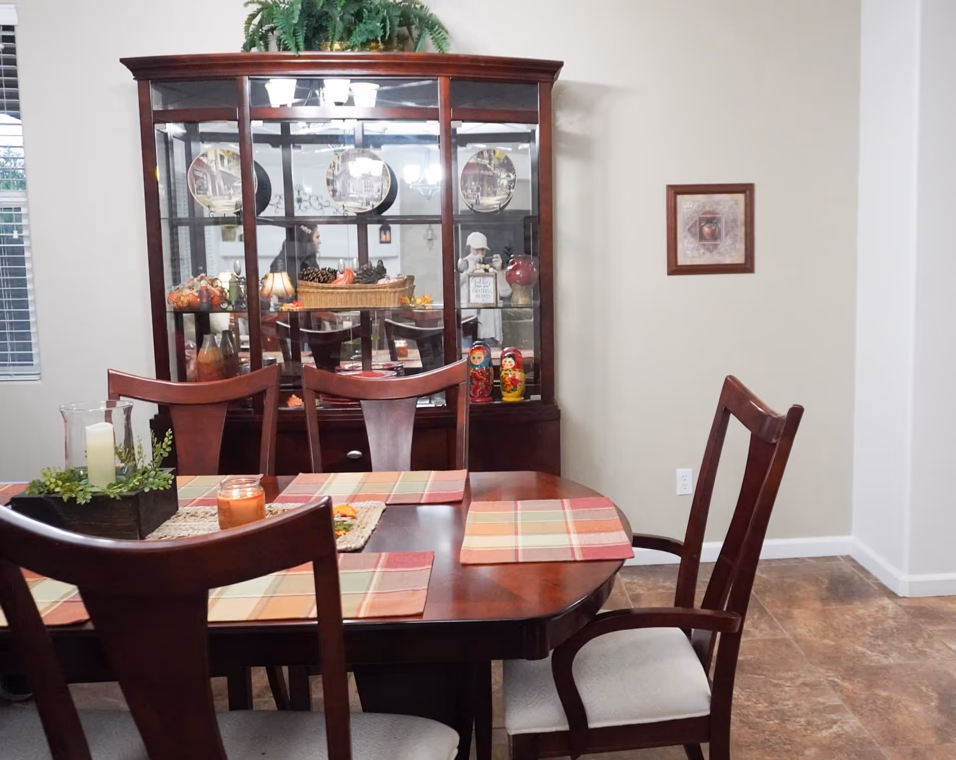 A dining room with a dark wooden table set with four chairs. The table has a centerpiece with a candle and greenery, and four plaid placemats. Behind the table is a large wooden china cabinet with glass doors displaying decorative plates and various ornaments. The room has light-colored walls, a framed picture on the wall, and a window with blinds on the left side.