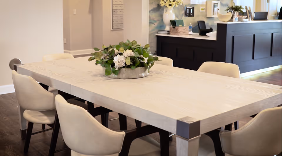 A modern dining area with a light wooden rectangular table surrounded by beige cushioned chairs. A decorative centerpiece with green leaves and white flowers is placed on the table. In the background, there is a dark wood reception desk with various items on top and a light-colored wall with artwork.