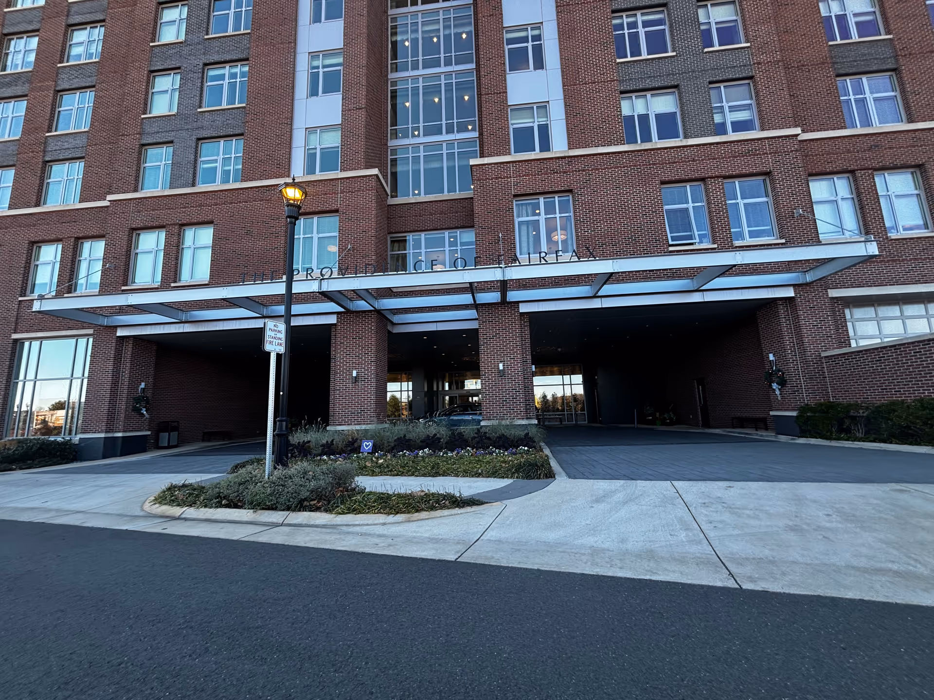 Front exterior view of The Providence Fairfax building showing a multi-story brick facade with numerous windows, a covered entrance with a glass canopy, a street lamp, and landscaped greenery in front.