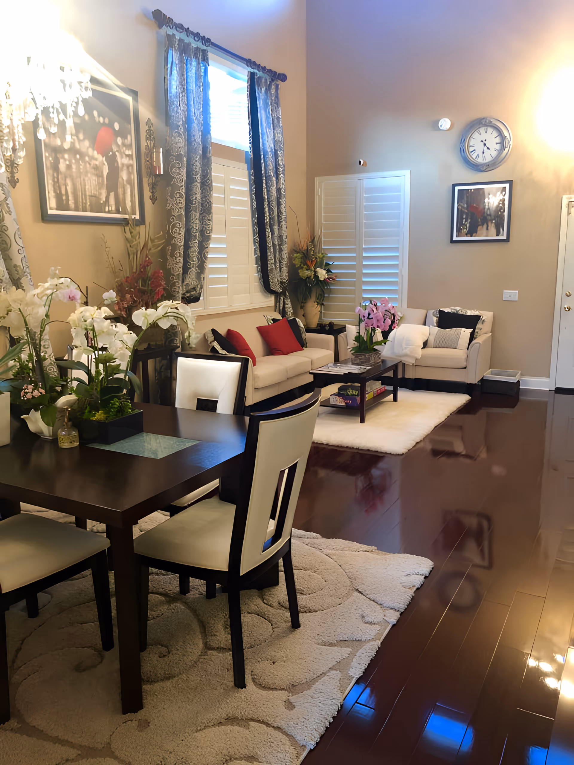 A cozy living and dining area with dark wooden floors and light-colored walls. The dining area features a dark wooden table with cream-colored chairs and floral centerpieces. The living area has a beige sofa and armchair with red and black cushions, a coffee table with flowers, and a white rug. The windows have white shutters and dark patterned curtains. Wall decorations include framed pictures and a clock.