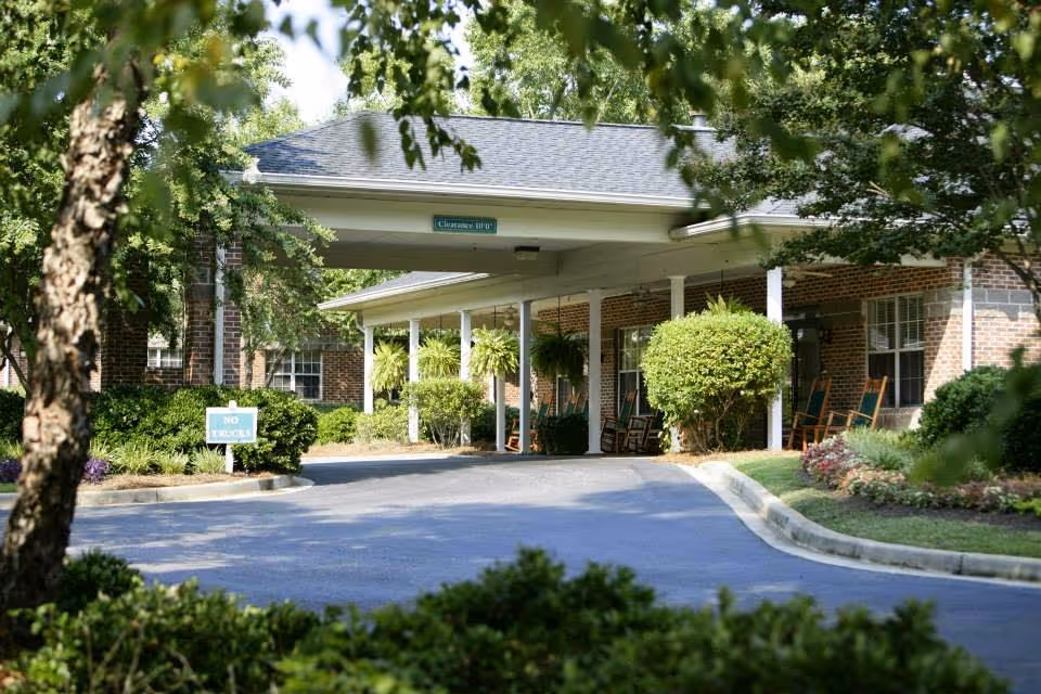 Entrance driveway of a senior living facility with a covered drop-off area supported by white columns. The building is made of brick and has several windows. There are green hanging plants and bushes along the walkway, with rocking chairs placed near the entrance. Trees and landscaped greenery surround the driveway.