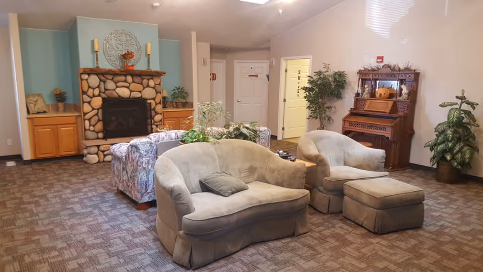 A cozy assisted living facility common area with a stone fireplace, two beige armchairs with a matching ottoman, a floral patterned sofa, and a wooden piano against the wall. The room has carpeted flooring, potted plants, and soft lighting.