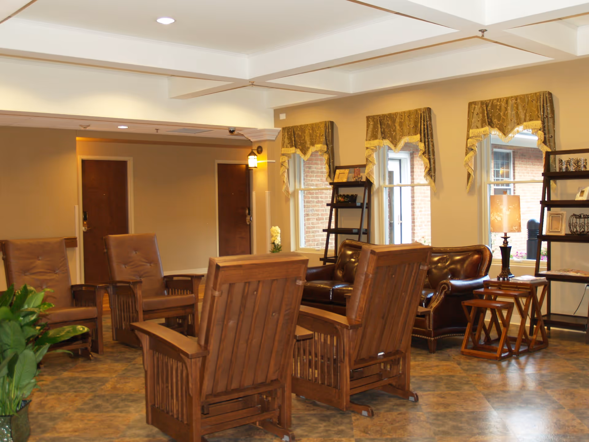A cozy sitting area in a retirement and assisted living facility featuring wooden armchairs with brown cushions, a dark brown leather sofa, small wooden side tables, a floor lamp, and decorative shelves near windows with valance curtains. The room has a tiled floor and beige walls with two closed wooden doors in the background.