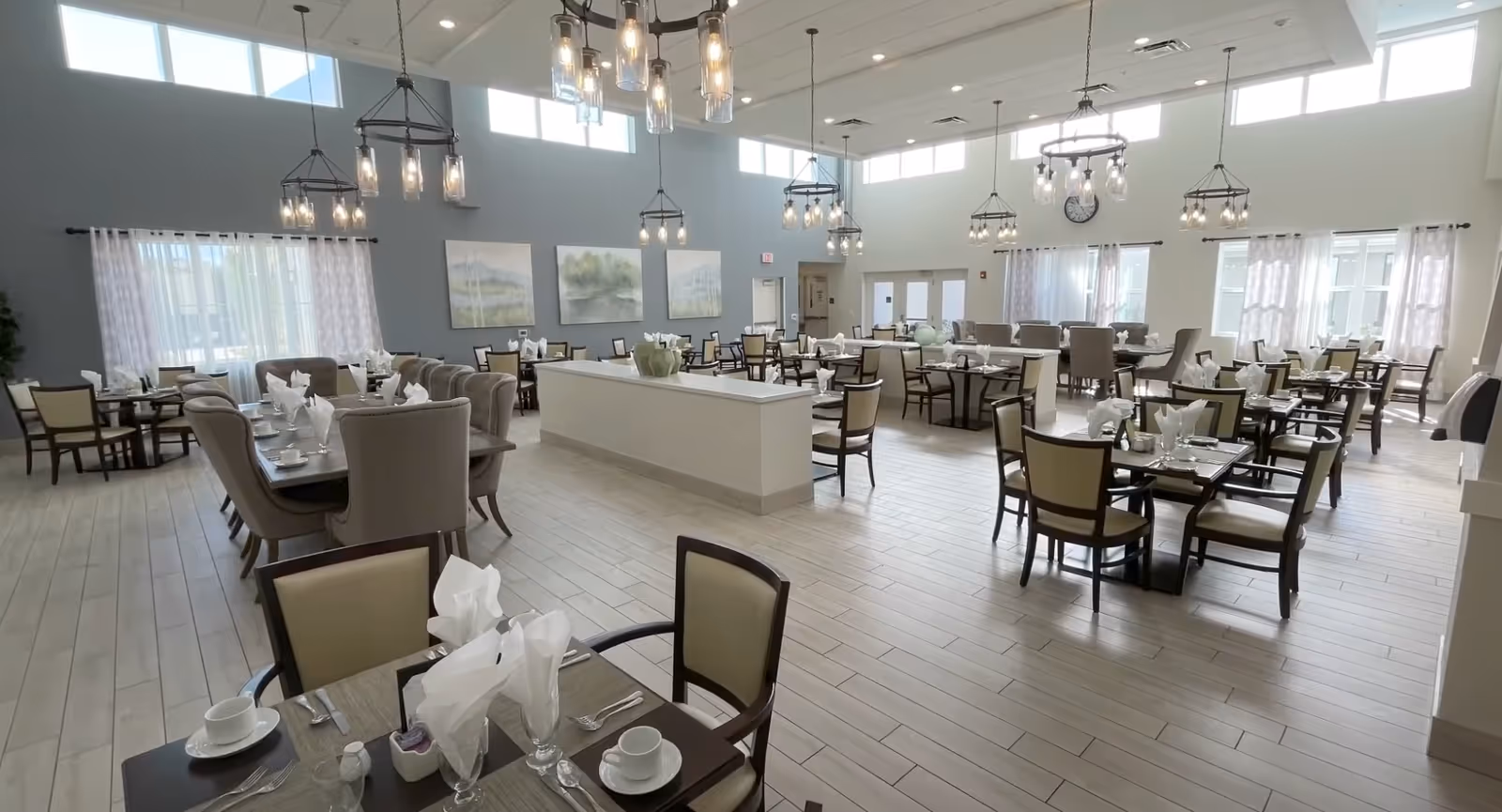 A spacious and well-lit dining room in a senior living community with multiple tables and chairs neatly arranged. Each table is set with white napkins, cups, and cutlery. The room features large windows with sheer curtains, modern hanging light fixtures, light-colored tiled flooring, and soft blue and white walls adorned with landscape paintings.