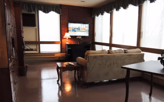 A cozy sitting area with a sofa, coffee table, lamp and wall-mounted TV in front of large curtained windows and a brick accent wall.