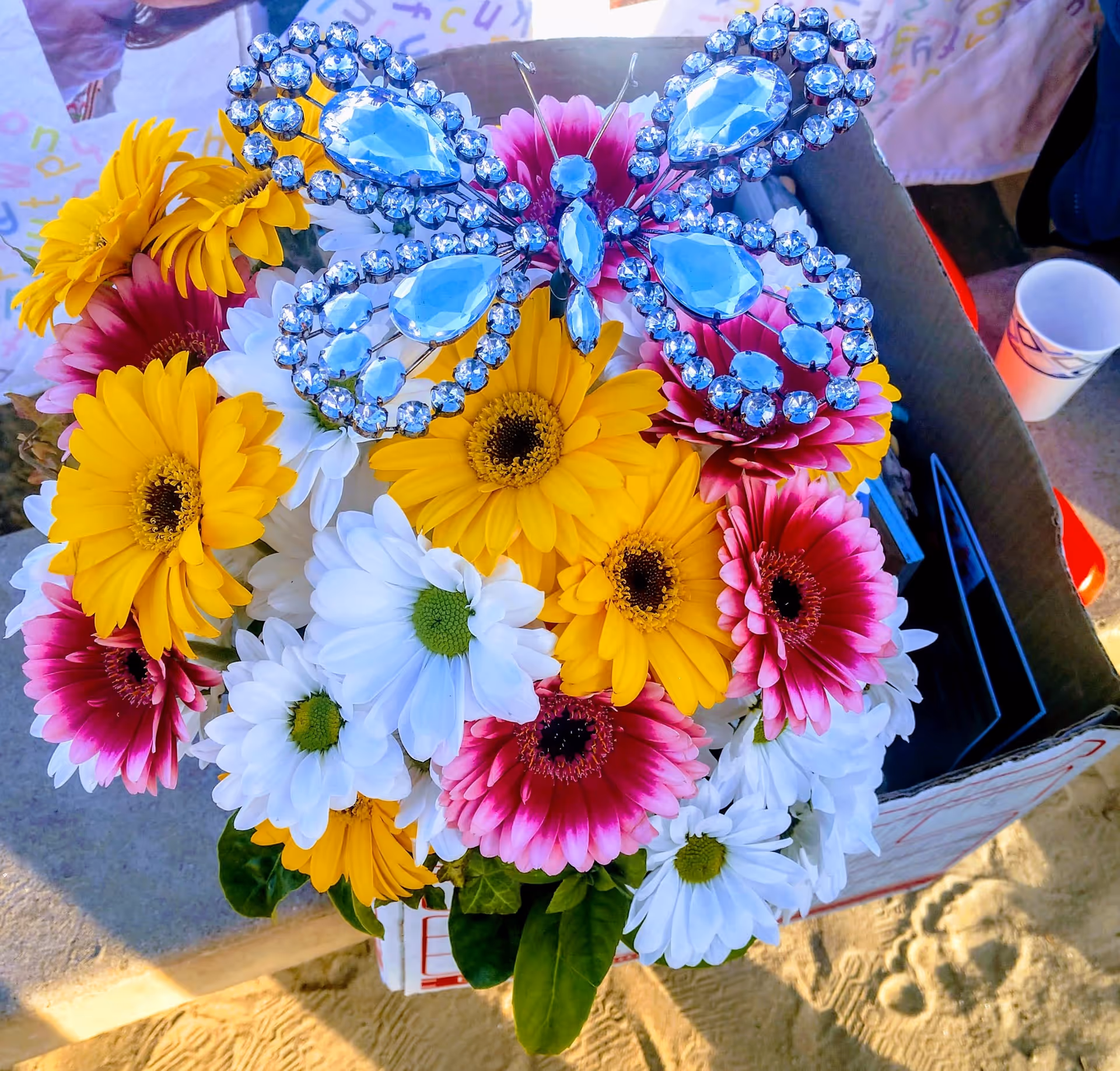 A colorful bouquet of yellow, white, and pink daisies topped with a blue jeweled butterfly ornament in a cardboard box.