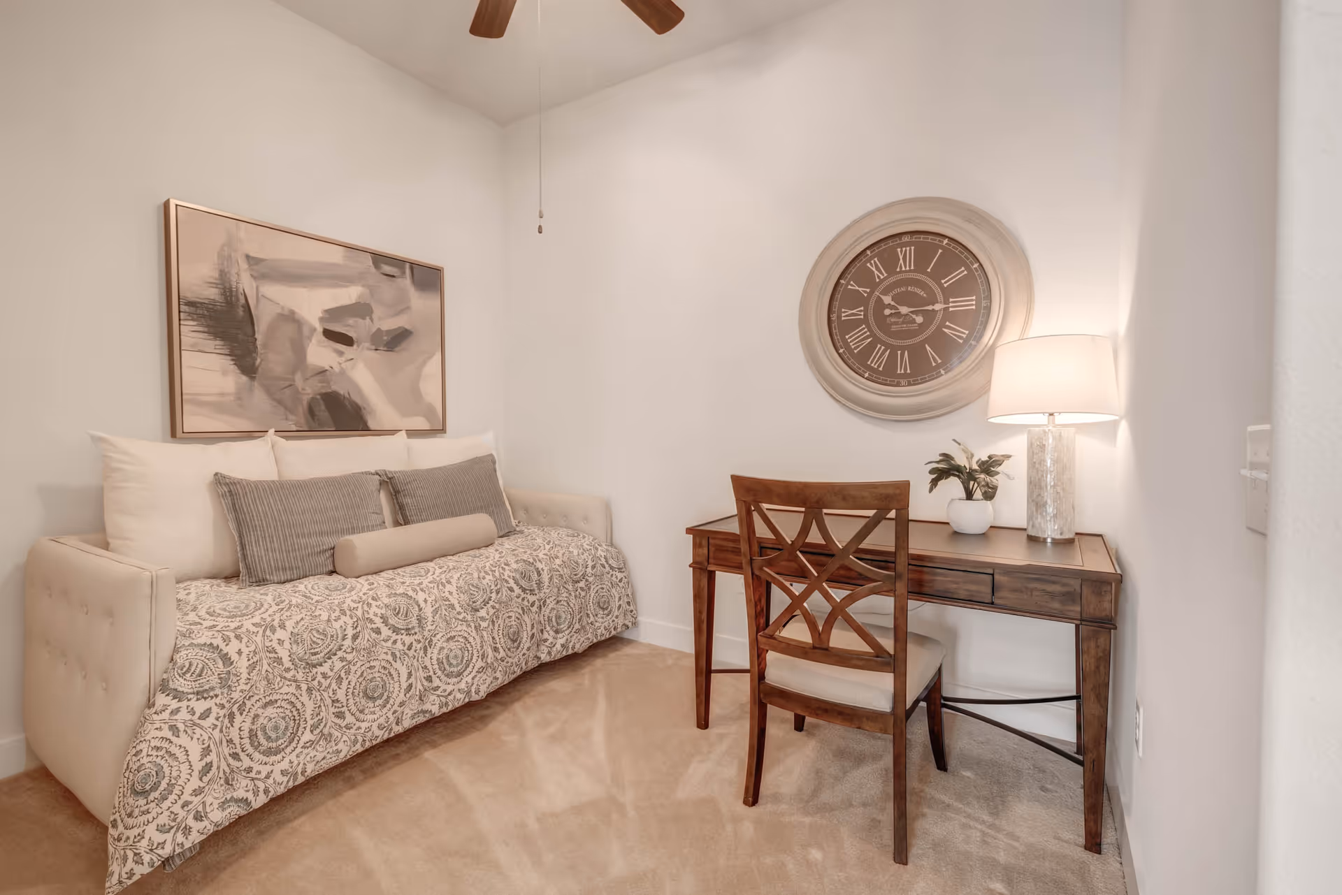 A cozy room with a beige daybed covered with patterned bedding and multiple pillows, a wooden desk with a matching chair, a table lamp, a small potted plant, and a large round wall clock above the desk. The walls are white and the floor is carpeted.